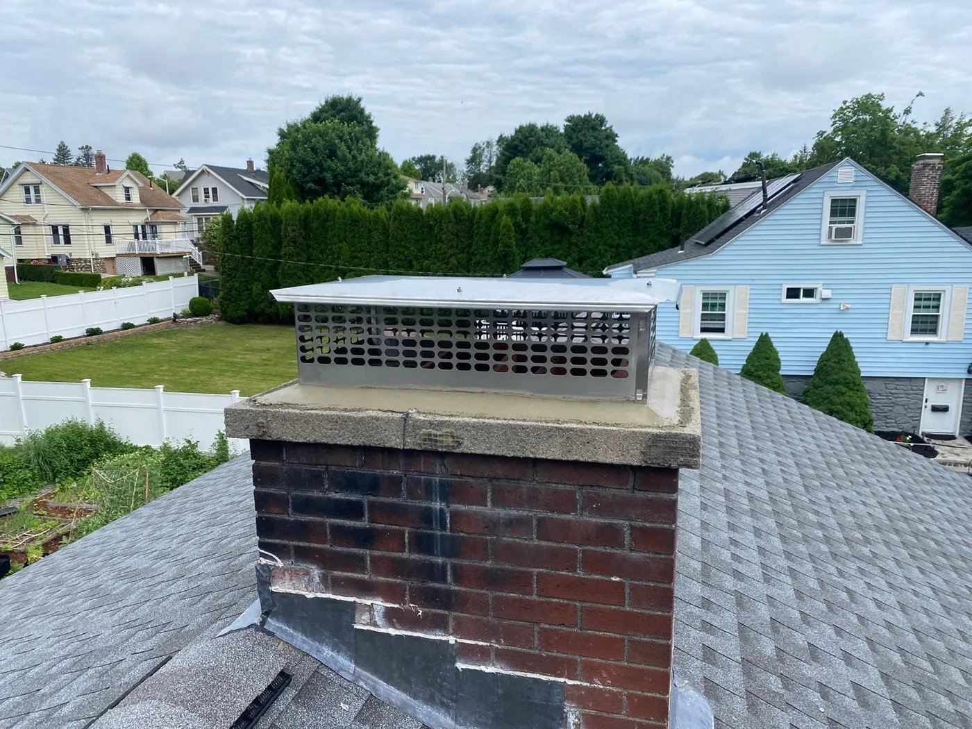 Rooftop view of a brick chimney with a new concrete crown and large stainless steel multi-flue cap, houses and trees in the background