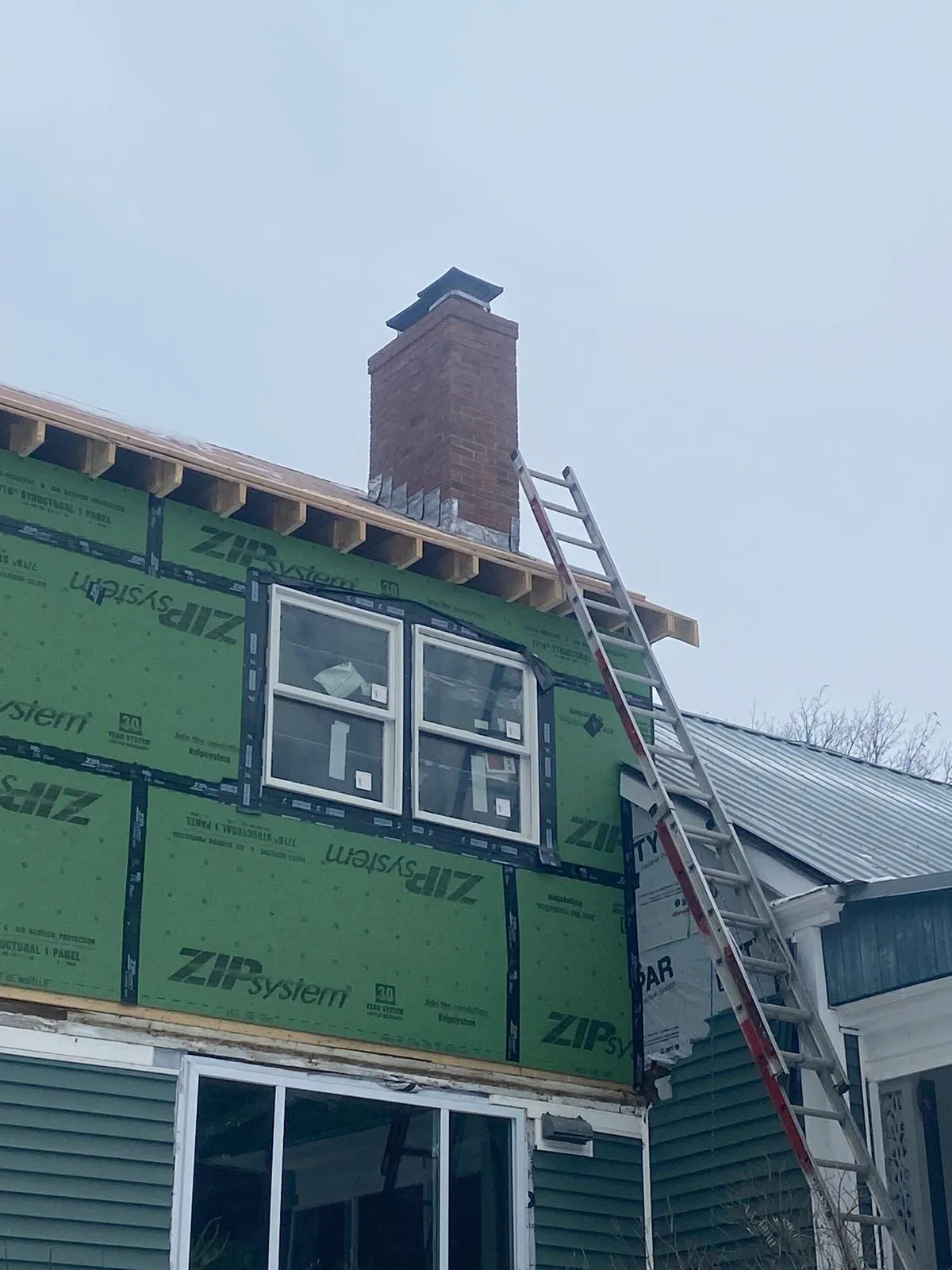 Brick chimney with new metal base flashing on a house under renovation with green sheathing panels and an extension ladder