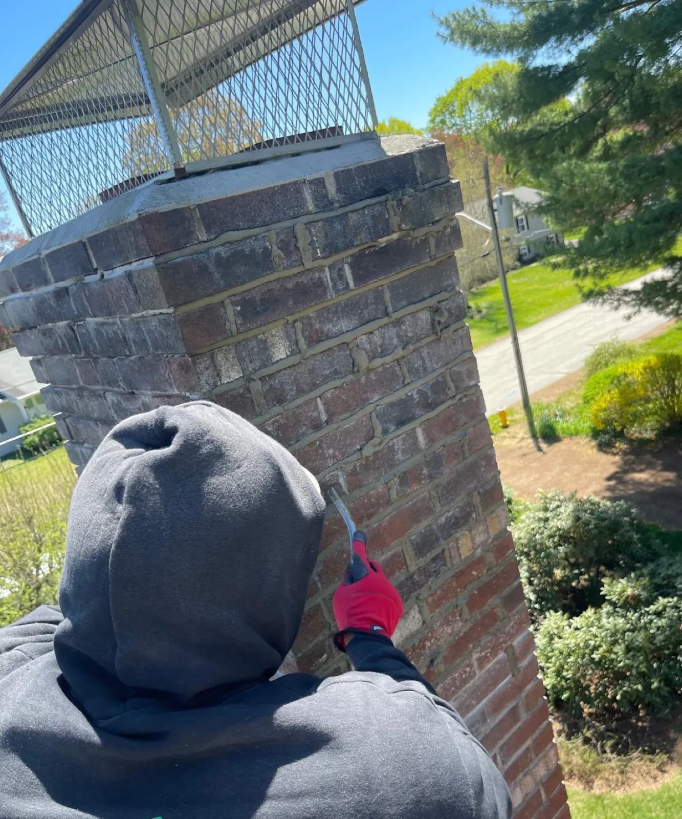 Worker in a hooded sweatshirt repointing mortar joints on a brick chimney from the rooftop