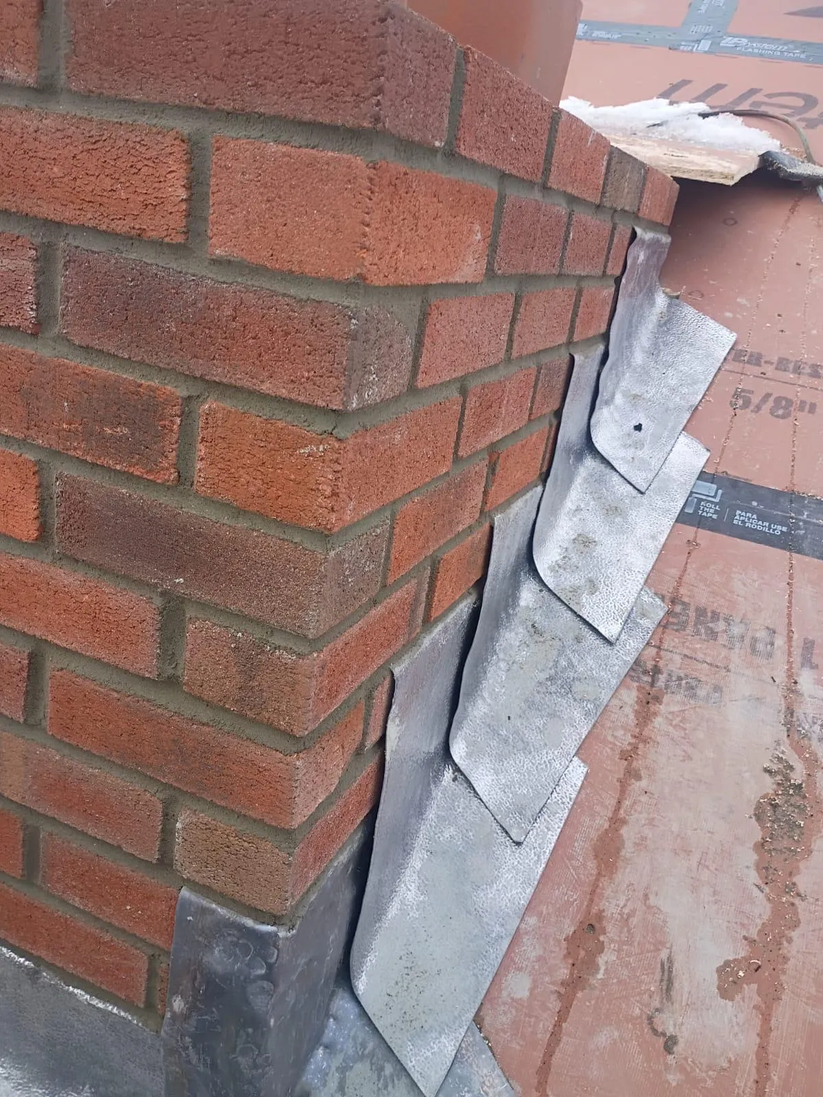 Lead step flashing installed along the corner of a newly built red brick chimney where it meets the roof deck