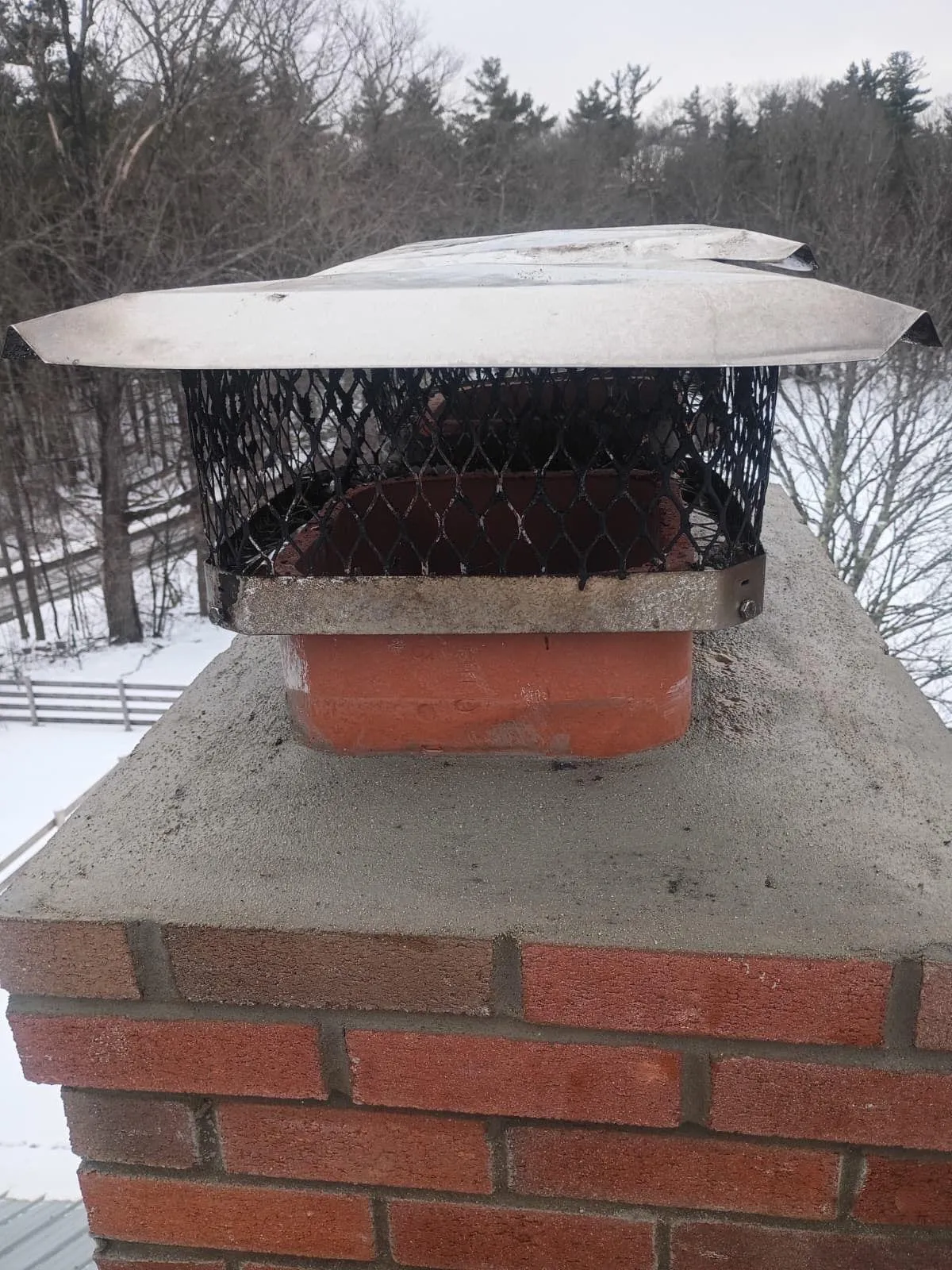 Stainless steel chimney cap with mesh sides installed on a clay flue tile atop a brick chimney, snowy trees in the background